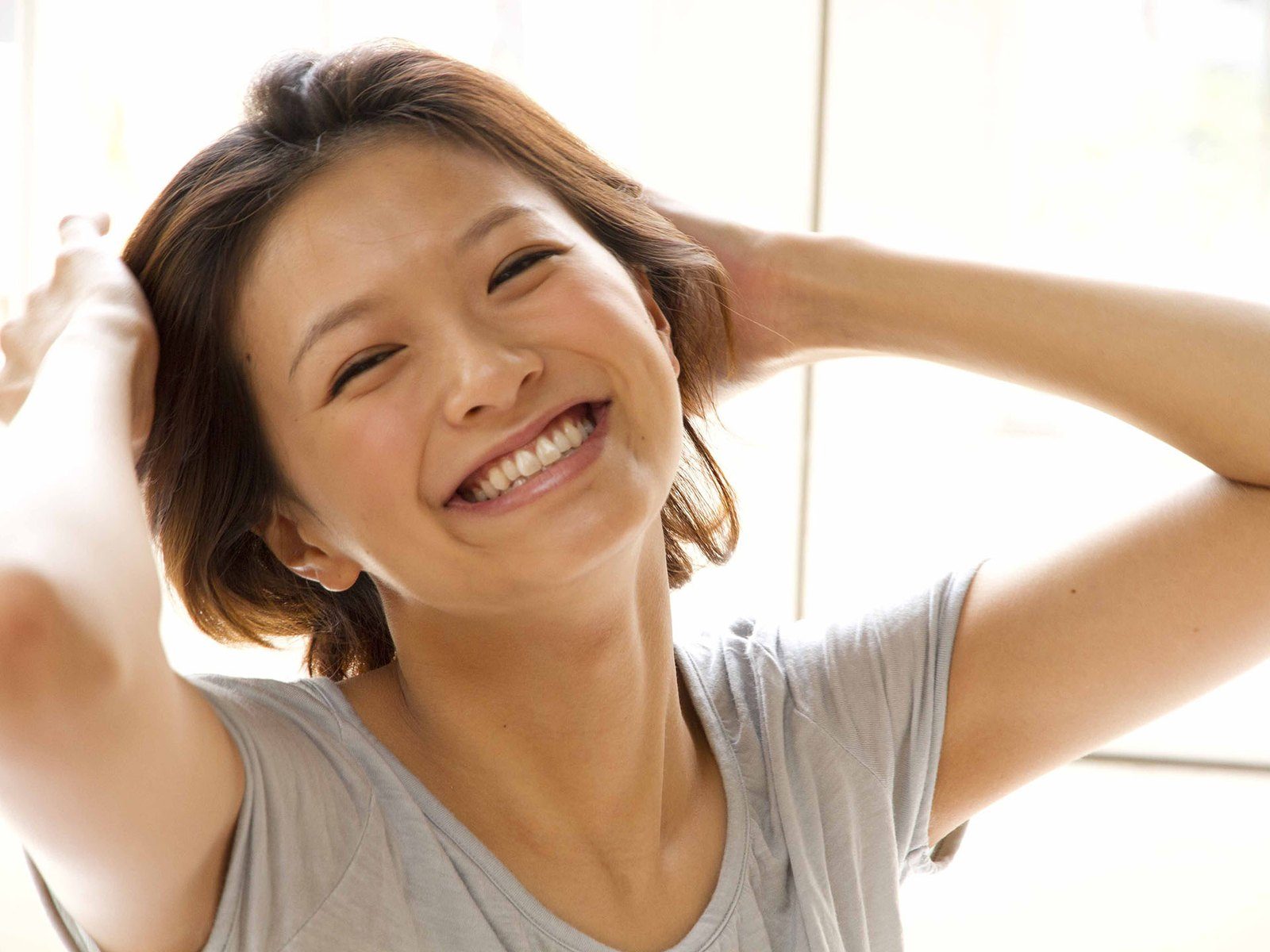 窓辺の光のなかで、両手を髪にあてて笑うショートヘアの女性